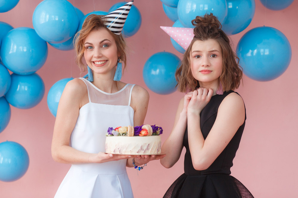 Two young women wearing party hats stand in front of a pink wall with blue balloons. One woman, smiling, holds a cake decorated with flowers. The other looks uncertain or surprised. Both are dressed in party attire.