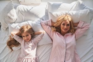 Una mujer y una niña de cabello largo y rubio están acostadas en una cama con almohadas y ropa de cama blancas, sonriendo y mirando hacia arriba, ambas vistiendo pijamas de rayas rosa claro, disfrutando de cuentos para dormir que fomentan la creatividad infantil.