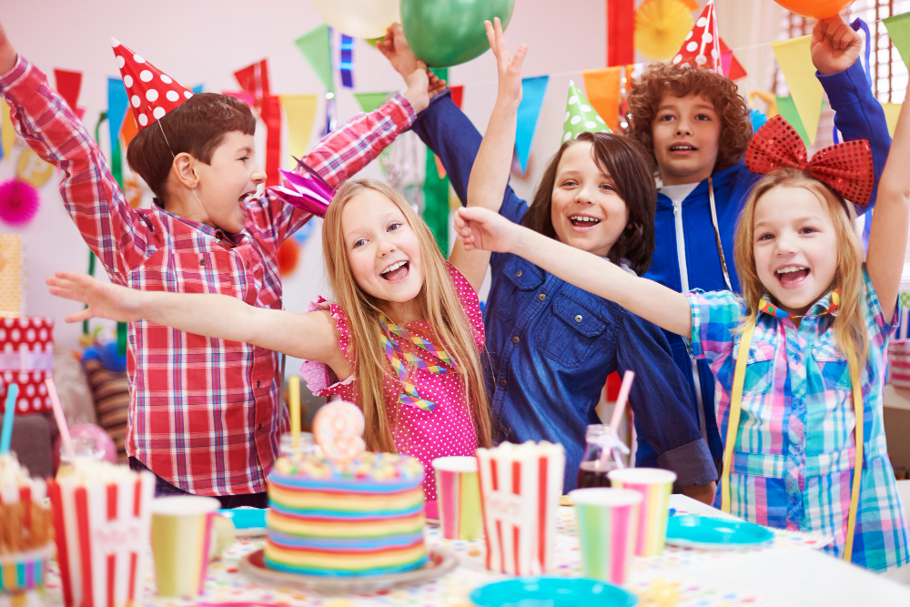Five children at a colorful birthday party, smiling and raising their arms in excitement. A decorated cake and snacks are on the table, with balloons and festive banners in the background.