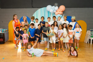 Un grupo de niños y dos adultos posan juntos en una fiesta de cumpleaños en un espacio cerrado. Los niños llevan ropa y calcetines coloridos, con globos y adornos de fiesta de fondo. Un niño yace juguetón en el suelo frente a ellos.