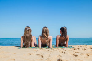 Three young women in swimsuits lie on mats at the beach, facing the sea. The sky is clear and blue, and the ocean is calm, creating a relaxed summer atmosphere.