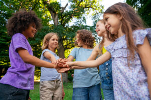 Five children stand in a circle outdoors, placing their hands together in the center, smiling and looking at each other. Trees and sunlight are visible in the background, creating a cheerful atmosphere.