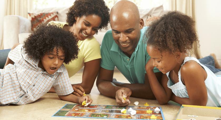 A family of four, including two children and two adults, lies on the floor happily playing a colorful board game together, smiling and engaged—just like when enjoying family friendly Switch games that everyone can play.