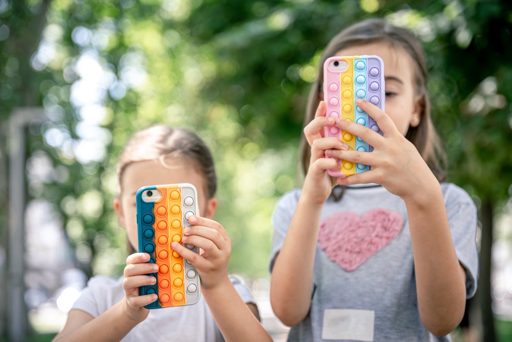 Dos chicas jóvenes al aire libre sostienen smartphones con carcasas de burbujas pop-it de colores que ocultan parcialmente sus rostros, tal vez por curiosidad sobre la verificación de edad en redes sociales. La vegetación borrosa del fondo sugiere que están en un parque o jardín.