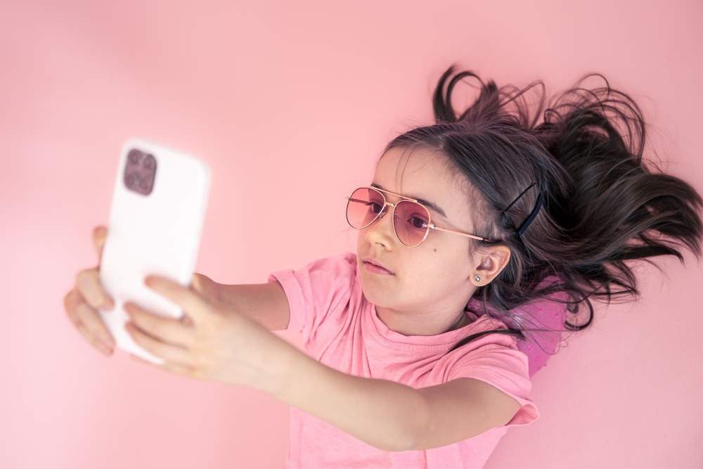 Una joven con gafas de sol rosas y camiseta rosa se tumba sobre una superficie rosa y sostiene un smartphone mientras se hace un selfie para las redes sociales. Su larga melena castaña se extiende alrededor de su cabeza.