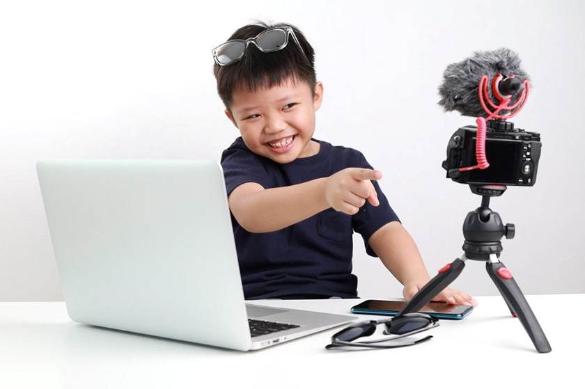 A smiling young boy sits at a desk with a laptop, smartphone, and sunglasses, pointing at a camera with a microphone on a tripod—capturing the excitement of kids content creation while learning about online safety.