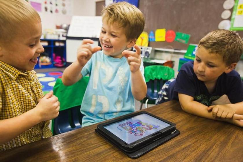 Three jóvenes creadores sit at a table in a classroom, smiling and laughing as they interact with a tablet displaying a colorful game. The background, decorated with educational materials, highlights the importance of seguridad online for niños.