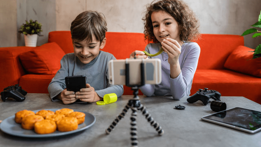 Dos niños sentados en una mesa gris frente a un sofá naranja se graban con un smartphone en un trípode, quizá para redes sociales con verificación de edad. Un niño sujeta el teléfono, el otro juega con un juguete. En la mesa hay magdalenas, mandos y una tableta.