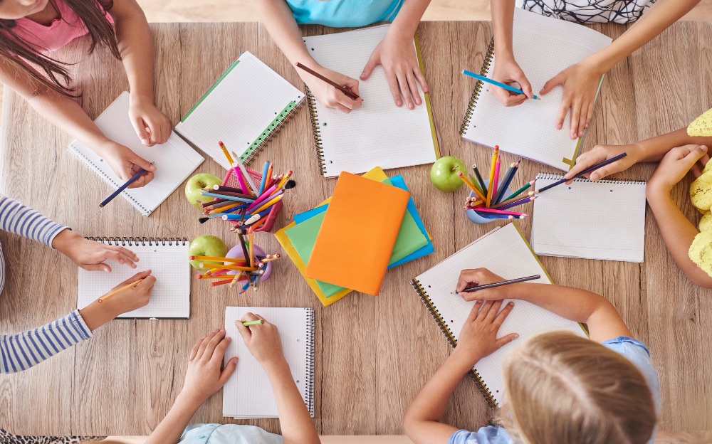Niños sentados alrededor de una mesa de madera escribiendo en cuadernos con lápices de colores. Sobre la mesa hay varios cuadernos, carpetas, manzanas y recipientes con lápices, que crean un ambiente alegre y creativo.