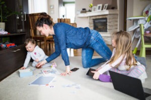 Una mujer arrodillada en la alfombra del salón juega con dos niñas. Una de ellas ordena las piezas de un puzzle y la otra sostiene una tableta. Hay juguetes y papeles esparcidos por el suelo, y la habitación parece acogedora y habitada.
