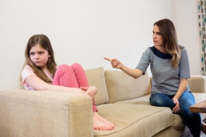Una mujer está sentada en un sofá, señalando y hablando con severidad a una joven que está de espaldas y parece disgustada, con las rodillas recogidas hasta el pecho.