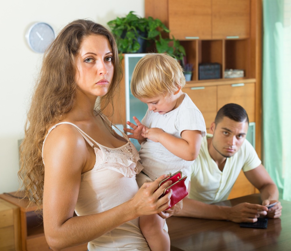 A woman holding a child and an open wallet stands in the foreground, looking serious, perhaps considering a parent checklist for the school year. In the background, a man sits at a table, also watching her. The setting appears to be a home.