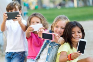Cuatro niños sonrientes sentados al aire libre tomándose selfies con sus smartphones, sosteniendo sus teléfonos delante de ellos y posando felices juntos en un día soleado en un parque.