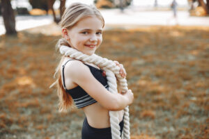 Una joven vestida con ropa deportiva sonríe a la cámara mientras sostiene una gruesa cuerda blanca sobre el hombro, de pie al aire libre sobre la hierba con árboles al fondo.