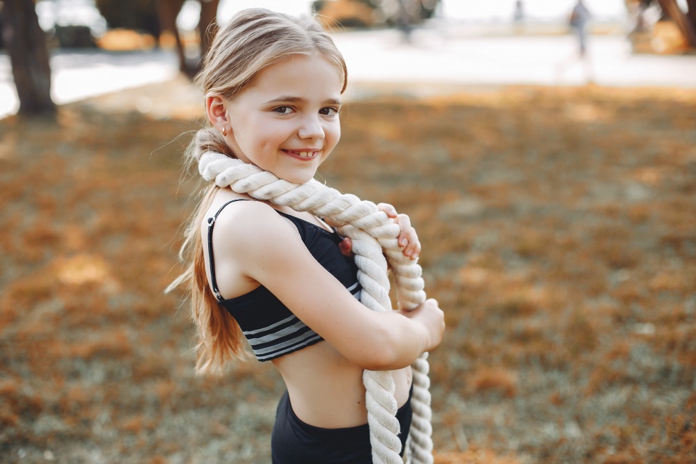 Una joven vestida con ropa deportiva sonríe a la cámara mientras sostiene una gruesa cuerda blanca sobre el hombro, de pie al aire libre sobre la hierba con árboles al fondo.