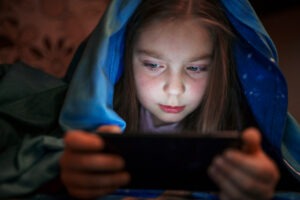 Молодая девушка с длинными каштановыми волосами сидит под голубым одеялом и сосредоточенно смотрит на смартфон, который она держит в обеих руках, ее лицо освещено светом экрана в тускло освещенной комнате.