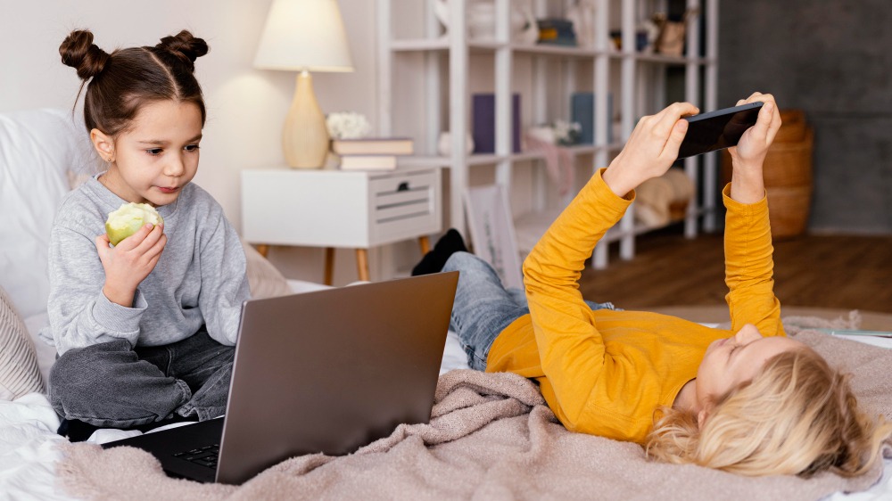 Двое детей отдыхают в уютной спальне: один сидит на кровати и ест яблоко, пользуясь ноутбуком, а другой лежит, держа над головой планшет. На заднем плане видны полки, книги и лампа.