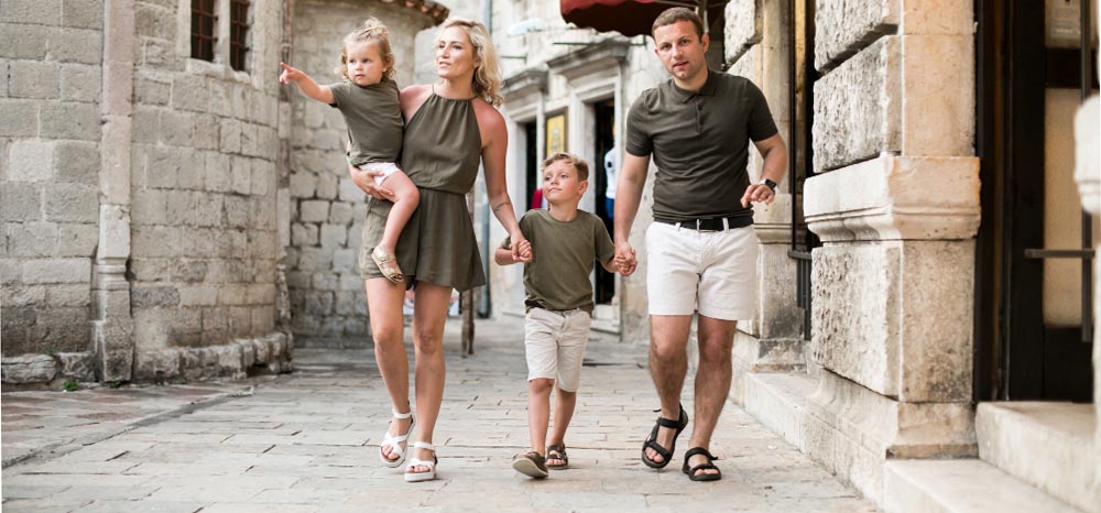 Una familia de cuatro miembros, vestidos con conjuntos verdes y neutros a juego, caminan juntos por una calle de piedra de una ciudad histórica. La madre lleva en brazos a una niña pequeña, mientras que el padre y el hijo caminan a su lado cogidos de la mano.