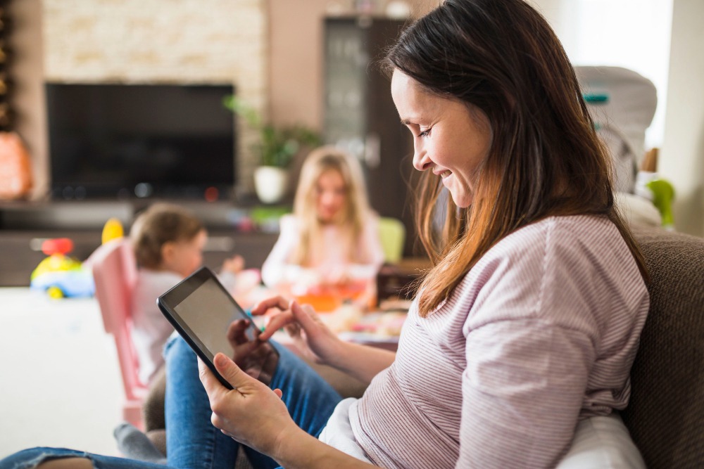A woman sits on a couch smiling while using a tablet, with two young children playing in the background—enjoying healthy habits for kids and fun indoor play Marbella style in a cozy living room.