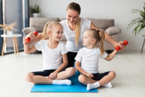 A woman sits on a yoga mat with two young girls, all in workout clothes. The smiling girls hold small orange dumbbells as the woman looks on, showing how Marbella indoor play and active play for children can make home workouts fun.
