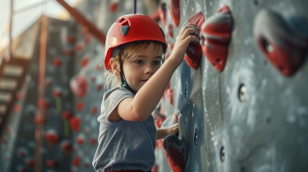 Un niño con casco rojo y camiseta gris trepa por una pared de roca agarrado a unas presas rojas y grises, con expresión concentrada. El fondo muestra más agarres y parte de la estructura de escalada.