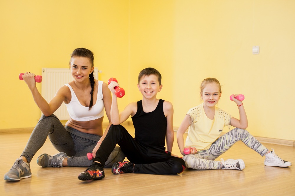 A woman, a boy, and a girl are sitting on a wooden floor in a gym in Marbella, smiling and holding small dumbbells. Dressed in sporty outfits, they're enjoying active play together in a bright yellow indoor play center.