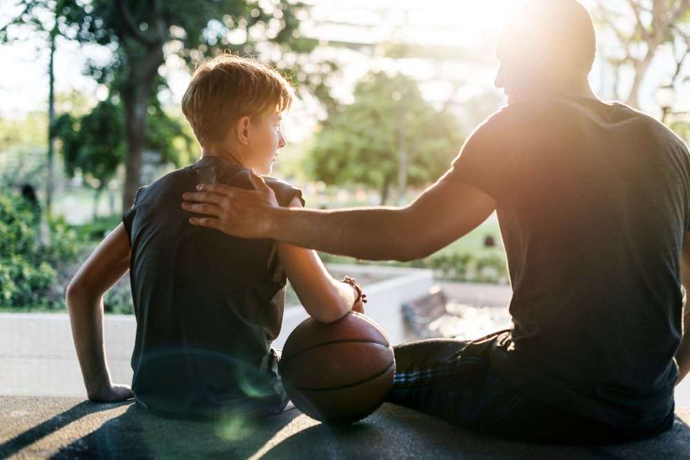 A sensitive child holding a basketball sits next to an adult, who has their arm around the child’s shoulders. They are outdoors on steps, with sunlight shining through trees—perfect for parenting tips and kids activities in Marbella.