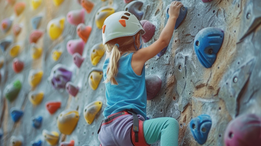 Маленький ребенок в шлеме и обвязке карабкается по крытой скальной стене с разноцветными креплениями, демонстрируя решительность и сосредоточенность. На заднем плане светит солнце, подчеркивая игривую атмосферу.