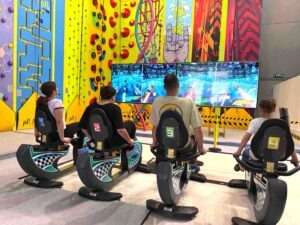 Four people sit on numbered motion simulator bikes, playing one of the best video games for kids 2026 on four large screens in a colorful, family-friendly arcade with bright walls and climbing structures—perfect for kids’ active play in Marbella.