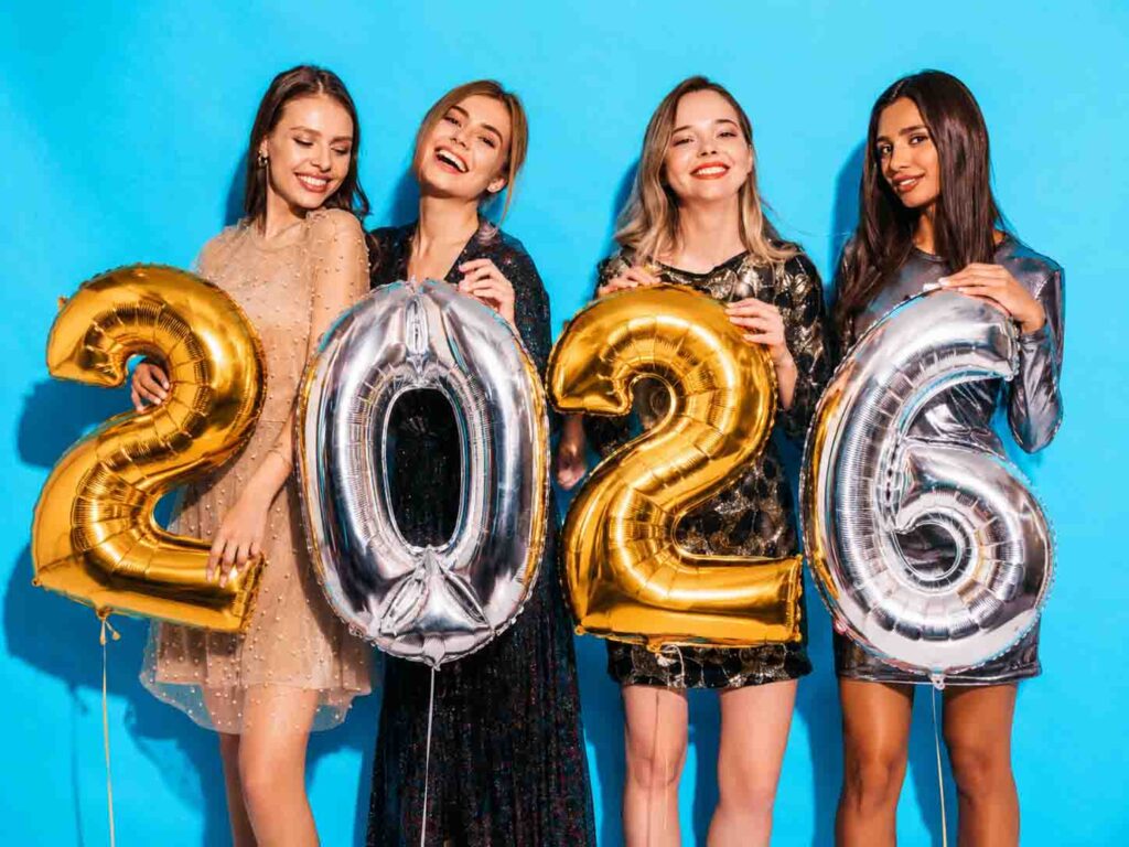 Four women stand in front of a blue background, smiling and holding large gold and silver balloons that spell out 2026, celebrating together after hosting an event focused on active play for kids.