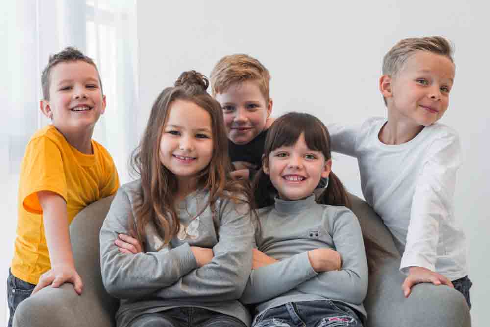 Cinco niños sonrientes están sentados muy juntos en un sillón gris sobre un fondo blanco liso, posando para la cámara con los brazos cruzados y atuendos informales con tops grises, blancos y amarillos.