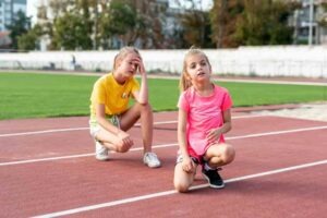 Dos chicas jóvenes vestidas con ropa deportiva se sientan y arrodillan en una pista de atletismo, con aspecto agotado después de hacer ejercicio. Una chica con camiseta amarilla apoya la mano en la cabeza y la otra, de rosa, se inclina hacia delante con la mano en la pierna.