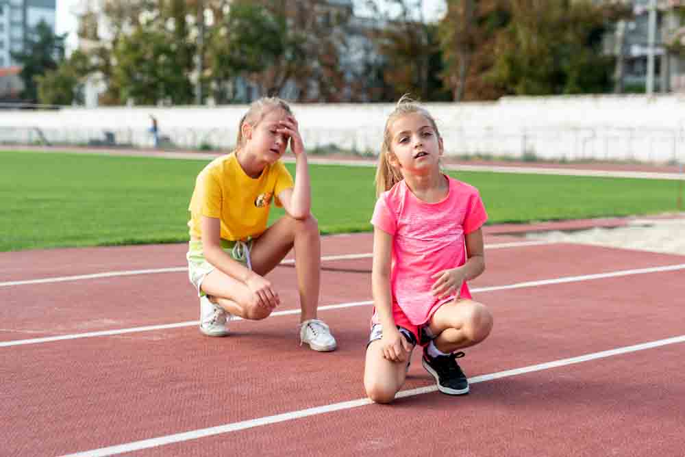 Dos chicas jóvenes vestidas con ropa deportiva se sientan y arrodillan en una pista de atletismo, con aspecto agotado después de hacer ejercicio. Una chica con camiseta amarilla apoya la mano en la cabeza y la otra, de rosa, se inclina hacia delante con la mano en la pierna.