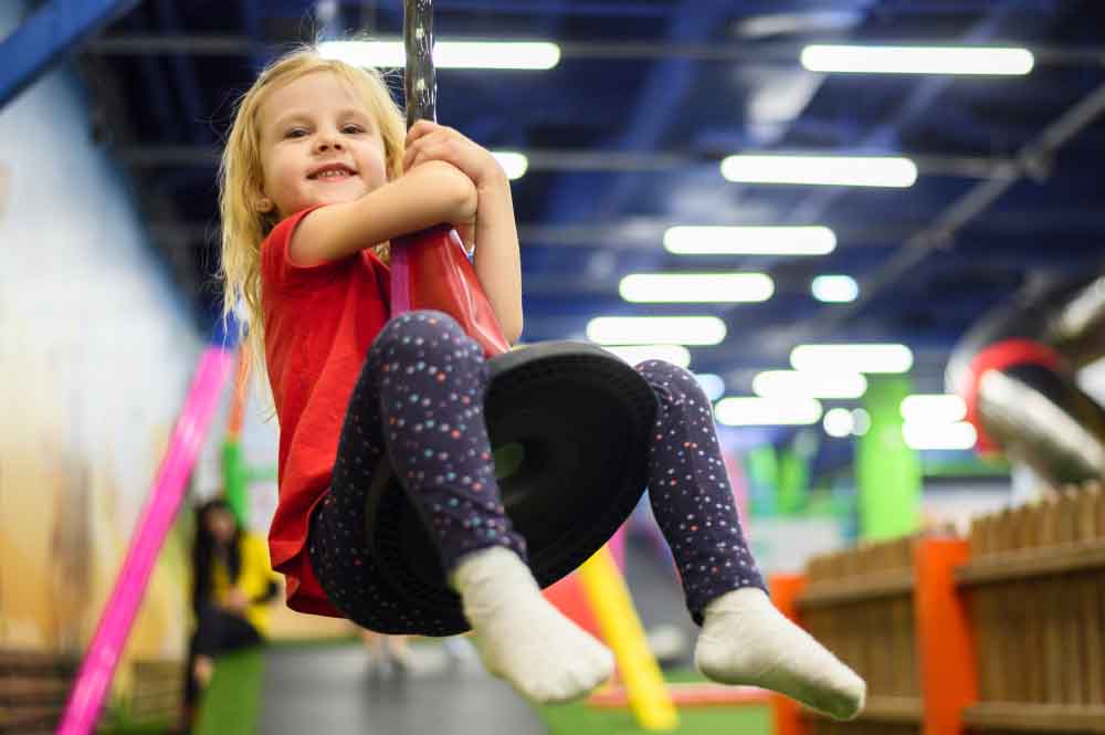 Una niña con una camiseta roja y leggings estampados sonríe mientras se balancea en un columpio de neumáticos en el interior, con juegos infantiles brillantes y fondo borroso.