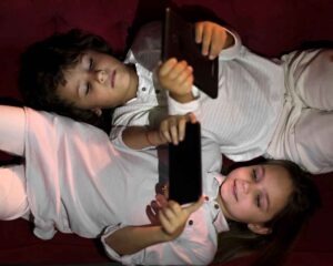 Dos niños vestidos de blanco están tumbados uno al lado del otro sobre una superficie roja, cada uno con una tableta encima, concentrados en sus pantallas.