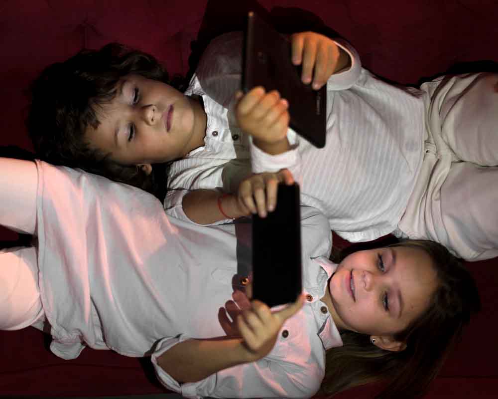 Dos niños vestidos de blanco están tumbados uno al lado del otro sobre una superficie roja, cada uno con una tableta encima, concentrados en sus pantallas.