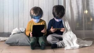 Two young children sit on the floor indoors, focused on digital devices. While they enjoy screen time, incorporating active play for kids—like games or movement breaks—beside their blanket and pillow can add balance to their day.