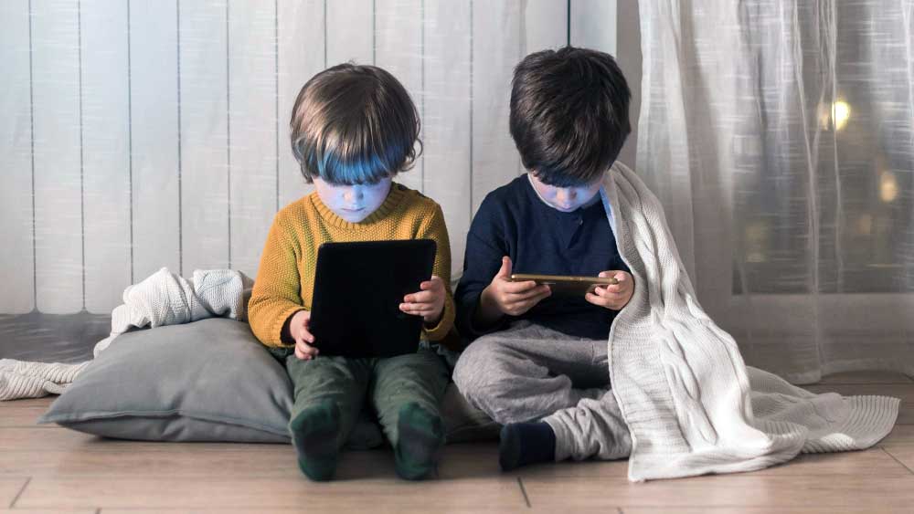 Two young children sit on the floor indoors, focused on digital devices. While they enjoy screen time, incorporating active play for kids—like games or movement breaks—beside their blanket and pillow can add balance to their day.