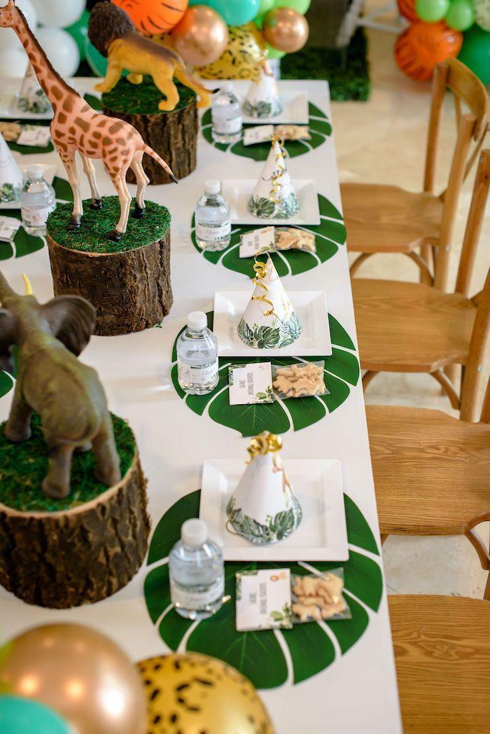 Una mesa de fiesta de temática selvática con figuras de animales, sombreros de fiesta, botellas de agua, manteles individuales con forma de hoja y galletas con forma de animales. Alrededor de la mesa hay sillas de madera y al fondo se ven globos decorativos.