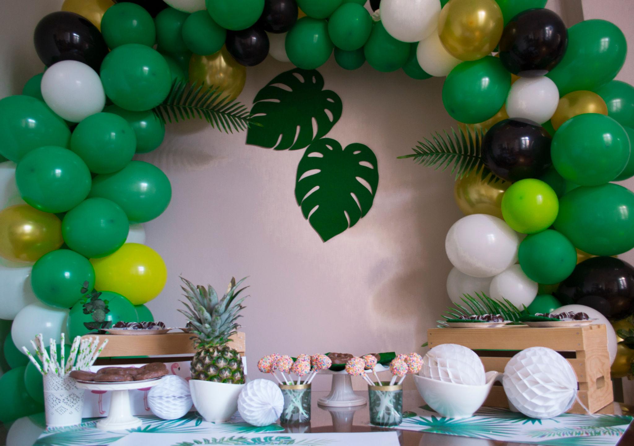 Una mesa de fiesta decorada con globos verdes, blancos, amarillos y negros formando un arco, hojas tropicales en la pared y varias golosinas, piñas y cuencos blancos sobre la mesa.