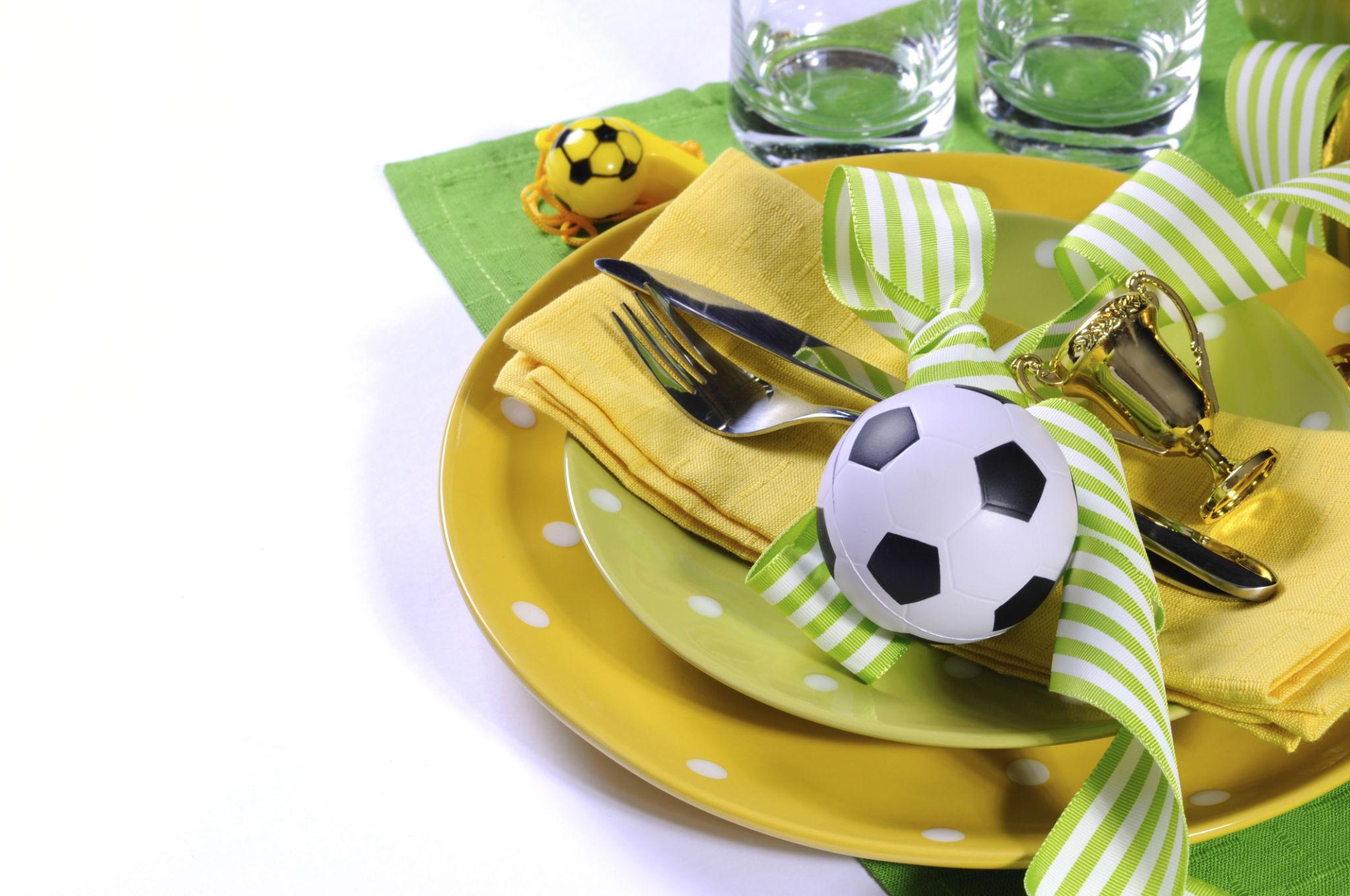 Servicio de mesa amarillo y verde con platos de lunares, dos vasos, servilletas amarillas y cubiertos. Decorado con un lazo, un pequeño balón de fútbol y un trofeo dorado en miniatura, sobre una servilleta verde.