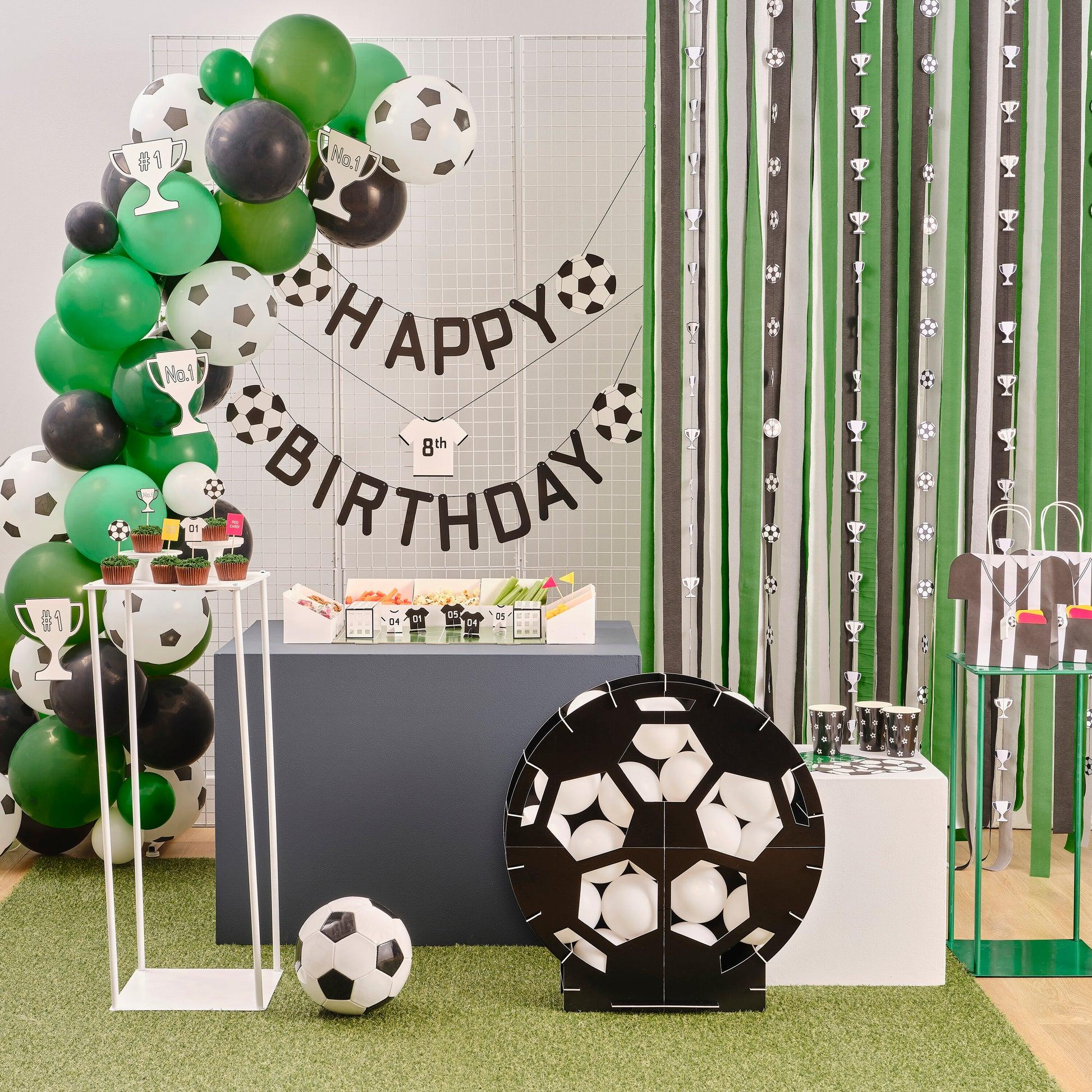 Fiesta de cumpleaños temática de fútbol con globos negros, blancos y verdes, una pancarta de Feliz Cumpleaños, decoraciones de balones de fútbol, aperitivos, magdalenas y recuerdos para la fiesta sobre césped artificial.