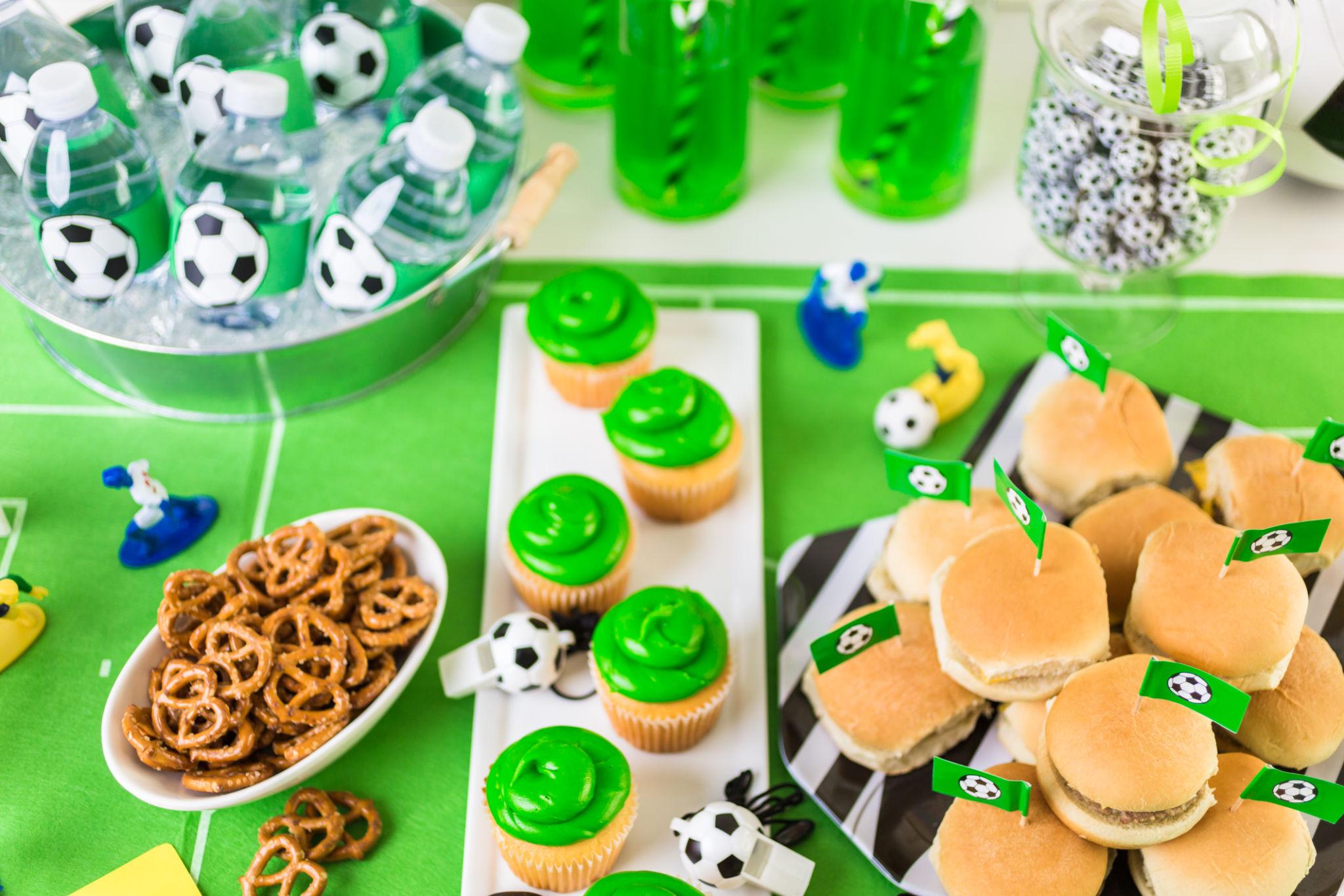 Una mesa de fiesta temática de fútbol con bebidas verdes, botellas de agua con tapones de balones de fútbol, magdalenas con glaseado verde, pretzels y pequeños bocadillos con banderines de fútbol sobre un mantel verde.