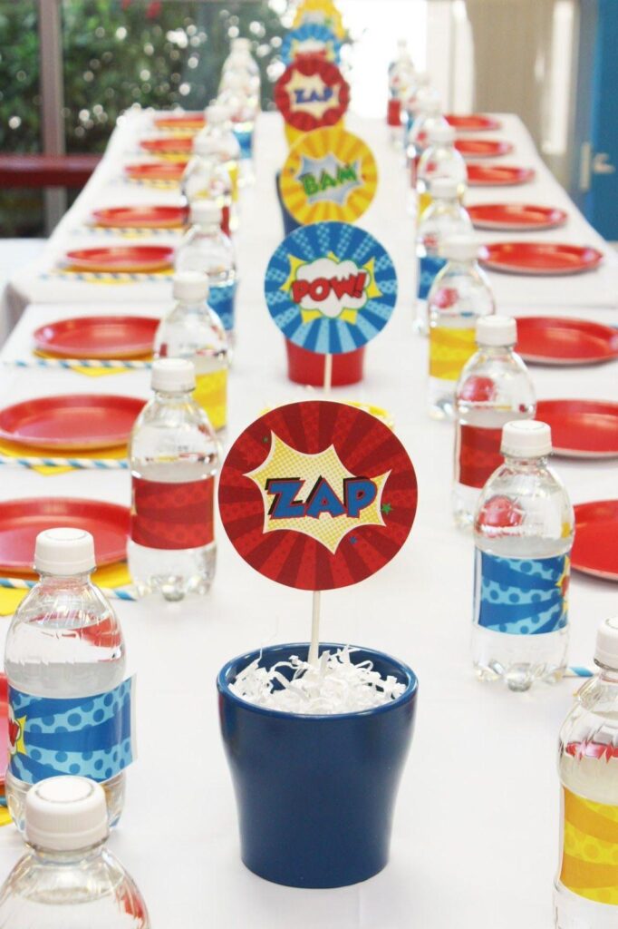 A long party table is set with red plates and bottles of water wrapped in superhero-themed labels, while centerpieces with comic book words like ZAP and POW add fun—perfect for discussing the AI effect on kids behavior at gatherings.
