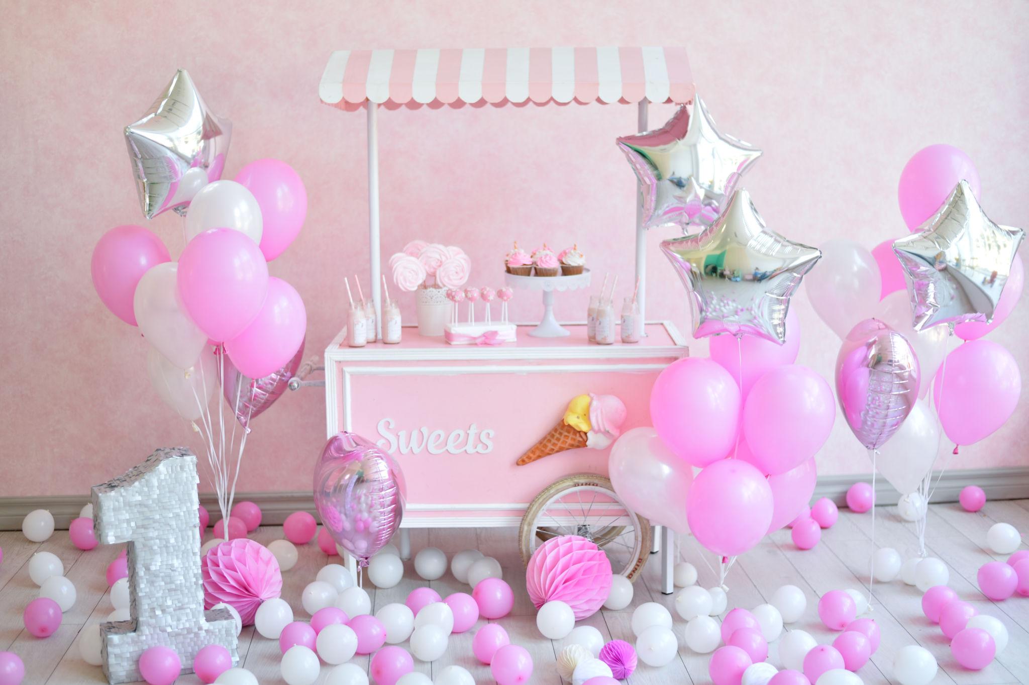 Carrito de caramelos rosas y blancos decorado con cupcakes, rosas rosas y tarros, rodeado de globos rosas, blancos y plateados, incluidos globos en forma de estrella y un gran número uno plateado entre bolas esparcidas por el suelo.