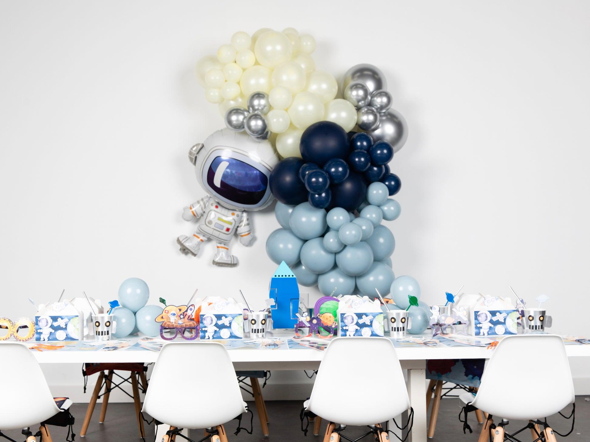 Una fiesta de temática espacial con sillas blancas alrededor de una mesa decorada con objetos de temática espacial y robótica. Detrás de la mesa, un gran globo de astronauta y globos azules, blancos y plateados colocados contra una pared blanca.