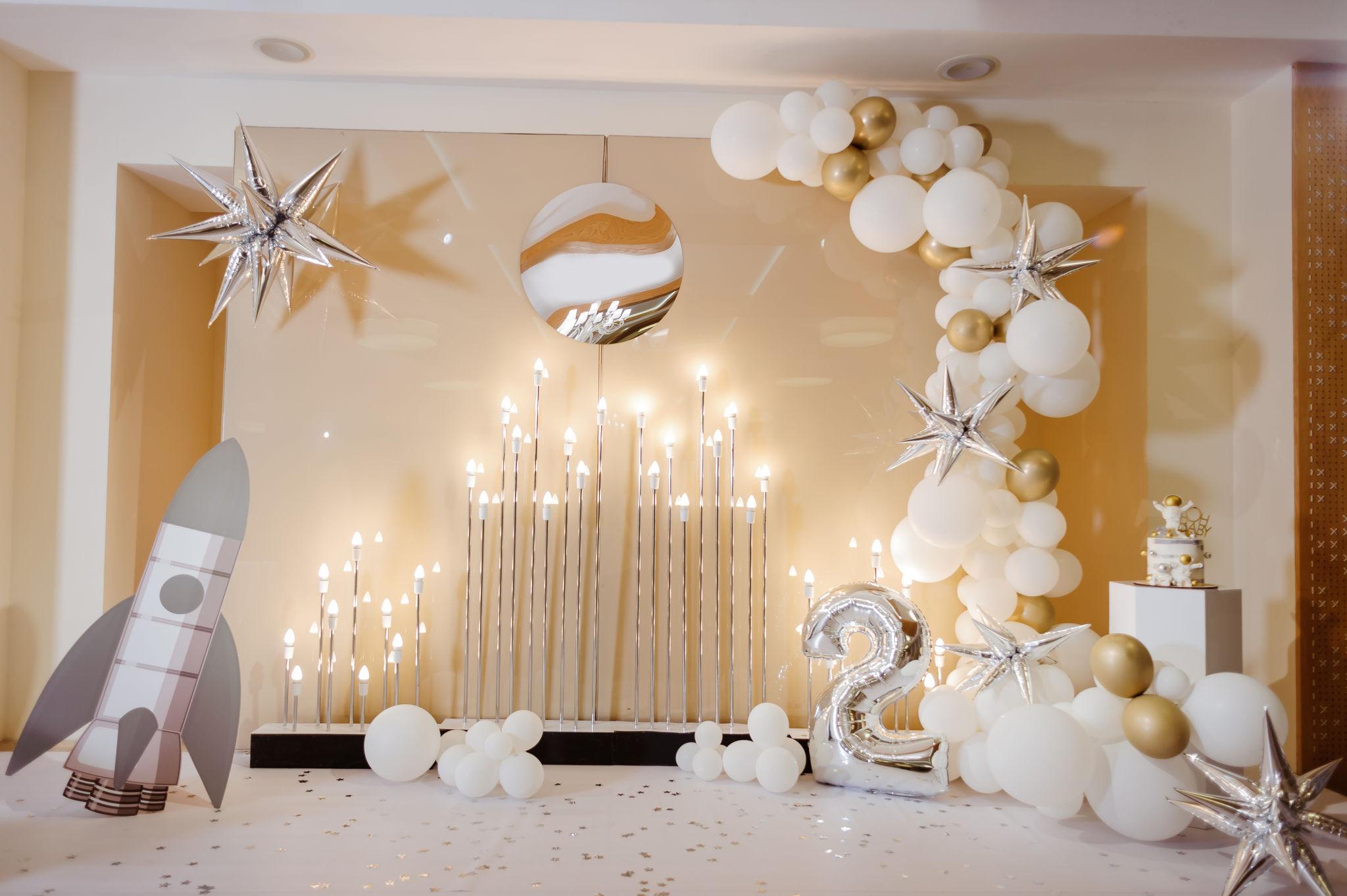 Montaje de cumpleaños de temática espacial con un cohete de cartón, globos blancos y dorados, globos con forma de estrella, una decoración de planeta, un gran globo plateado 2 y una tarta sobre un pedestal. En el fondo hay velas encendidas.