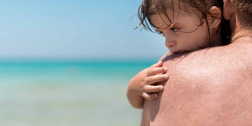 A young child with wet hair rests on an adult’s bare shoulder, looking thoughtfully into the distance. The calm blue sea and sky evoke a peaceful beach setting, perfect for creating cherished birthday memories for kids.