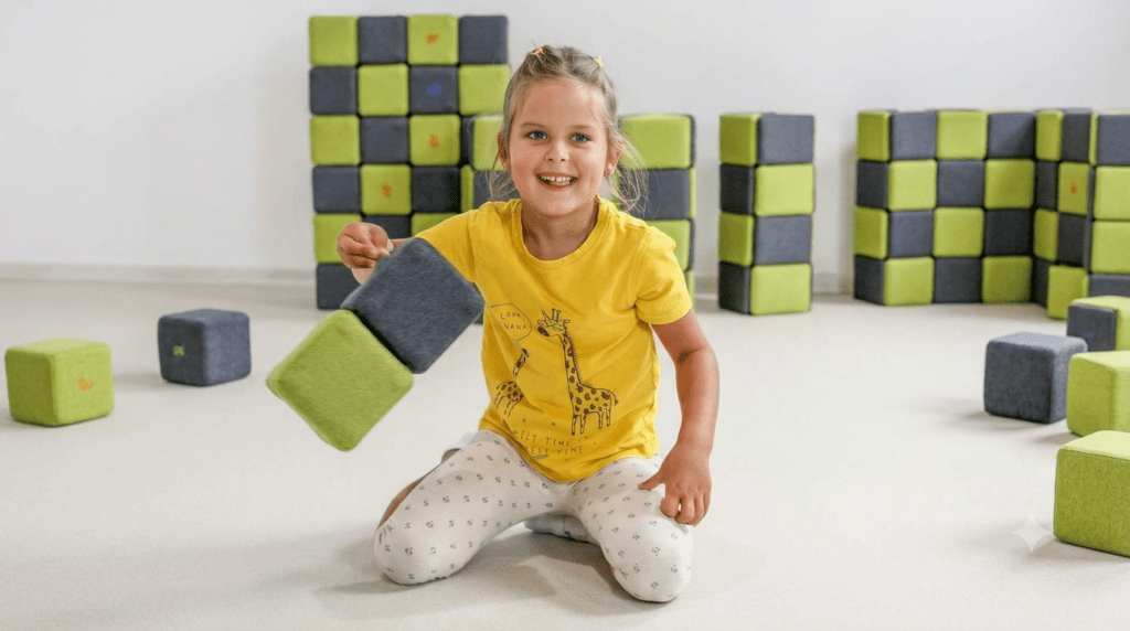 Una niña pequeña está sentada en el suelo, sonriendo, y sostiene dos bloques blandos. Lleva una camiseta amarilla con una jirafa y unos leggings estampados. Al fondo se ven bloques apilados de color verde y gris.